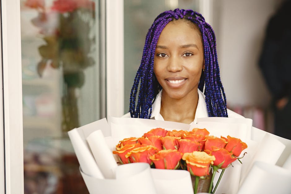 A young woman with dark skin and long, braided purple hair is holding a bouquet of fresh roses wrapped in white paper. The roses are a vibrant mix of coral pink and orange hues, arranged in a compact,