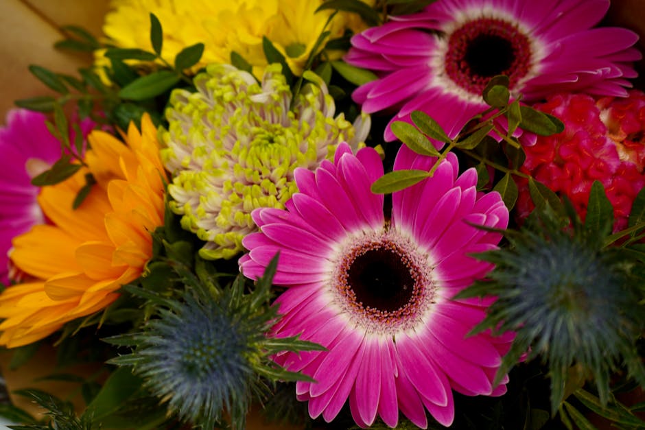A vibrant floral arrangement featuring large pink daisies with dark centers, surrounded by bright yellow sunflowers, pale green chrysanthemums with tightly packed petals, spiky blue thistles, and lush