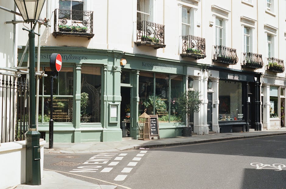 A storefront of a flower shop with a pale green facade, large display windows showcasing various plants and floral arrangements, and a small chalkboard sign outside. The shop is situated on a corner o
