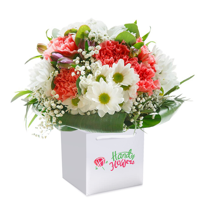 Set against a pristine, white background, this exuberant floral arrangement stands upright in a modern white square gift bag-a testament to the refined artistry found in Mitcham floristry. At its heart, lush coral-pink and vibrant red carnations cascade in tightly packed layers, their signature ruffled petals creating a velvety, inviting texture evocative of the seasonal blooms found near Mitcham Eastfields. Emerging between these passionate tones, pristine white daisies beam with golden-green centers, their slender petals radiating a sense of youthful optimism and gentle cheer. Wisps of airy baby's breath meander through the bouquet, their tiny white blossoms providing a delicate counterpoint and soft, feathery texture amid the more robust blooms. Subtle hints of unopened, purple-edged buds introduce quiet sophistication and depth, suggesting the unfolding of future moments to cherish. A collar of deep, glossy green foliage wraps protectively around the base, amplifying the sense of freshness and structure, much like the natural green havens dotted across the local area. The white gift bag, accentuated by sturdy ribbon handles, lends the arrangement an air of understated elegance and ease-a thoughtful detail for those seeking doorstep delivery to friends on London Road or neighbours near Mitcham Junction. Each bloom seems thoughtfully placed, the palette both vibrant and harmonious, making this bouquet a beautiful expression of joy, celebration, or simply a heartfelt gesture to brighten someone's day in the Mitcham community.