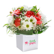 A delightful snapshot of spring, this vibrant bouquet effortlessly exudes warmth and cheer from its bright perch inside a pristine white gift bag-an arrangement echoing the spirit of Mitcham's cheerful neighbourhoods. The eye is first drawn to a lush gathering of soft coral and red carnations, their ruffled, luxurious petals packed tightly together to create a feeling of abundance and plushness. Mingling beside them, crisp white daisies with lively lime-green centers add a fresh brightness, their playful energy reminiscent of sunny days at Grove Park. Sprigs of delicate white baby's breath thread through the display, their airy blossoms bringing a sense of innocence and gentle movement. On one edge, small purple-tinged buds offer a whisper of hidden promise and elegant mystery. All are enfolded by swirling arcs of glossy green leaves that wrap around the arrangement, lending both visual grounding and texture. The gift bag itself is broad and modern, topped by neat ribbon handles for easy gifting or delivery-a familiar comfort in the Mitcham area, where thoughtful gestures travel from door to door. Bathed in soft, diffuse light, each stem seems to radiate freshness and care, making this bouquet an ideal choice for celebrating birthdays, congratulating achievements, or sharing joy within the vibrant community. Crafted with love by local florists, this arrangement captures the uplifting spirit and beauty found right here in CR4.